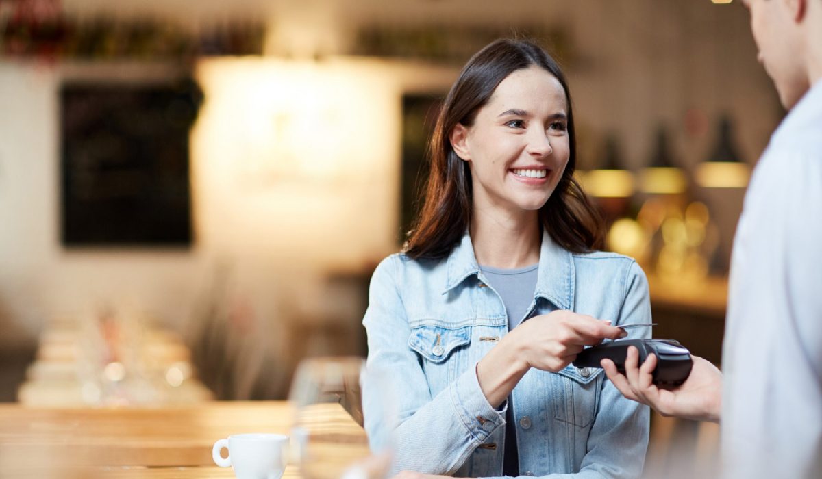 Young smiling woman with card holding it over payment terminal while paying for her order in cafe